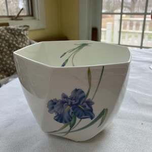 May include: A white, octagonal ceramic bowl with a blue iris floral design. The bowl has a glossy finish and is set on a white tablecloth. The iris is depicted with blue petals and green leaves.