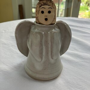 May include: A ceramic angel figurine with a surprised expression. The angel has a light-colored body, wings, and head with detailed facial features. A small, round, brown piece sits atop the head, with a wick extending from it.