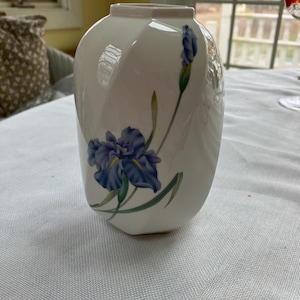 May include: White ceramic vase with a stylized blue iris flower and bud design. The vase has a unique, curved shape and a slightly flared top. The iris has blue petals and green leaves.
