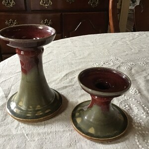May include: Two ceramic candle holders with a green and red glaze. The larger holder has a wider base and a taller stem. The smaller holder has a smaller base and a shorter stem.