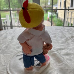 May include: A handmade doll wearing a yellow cap with a red propeller, a white shirt, blue jeans, and holding a brown object. The doll is standing on a white oval base.
