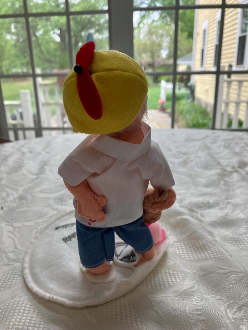 May include: A handmade doll wearing a yellow cap with a red propeller, a white shirt, blue jeans, and holding a brown object. The doll is standing on a white oval base.
