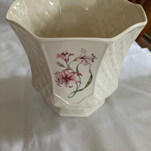 May include: Cream-colored ceramic planter with a woven basket design. The planter features a floral design of pink lilies. The planter has a hexagonal base and a scalloped rim.