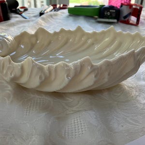 May include: Cream-colored, shell-shaped decorative bowl. The bowl has a textured, wavy design along the edges and a smooth interior. The bowl features a spiral design on one end, adding a unique touch to the overall aesthetic. This decorative bowl is perfect for displaying small items.