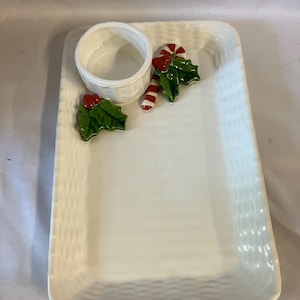 May include: A rectangular white ceramic serving tray with a woven basket-weave texture. The tray features an attached small round bowl, decorated with sculpted green holly leaves, red berries, and a red and white striped candy cane accent.