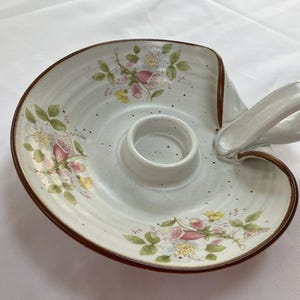 May include: Cream-colored ceramic candle holder with a handle and floral design. The holder features a central ring for the candle and is decorated with pink, yellow, and white flowers and green leaves. The rim is a dark brown color.