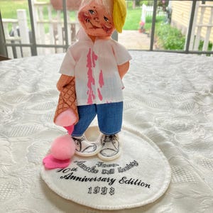 May include: A vintage doll with a yellow hat and a white shirt stained with pink. The doll is wearing blue jeans and white sneakers. It is holding a brown ice cream cone and standing on a white base with text that reads "Timothy Annalee Doll Society 10th Anniversary Edition 1993."
