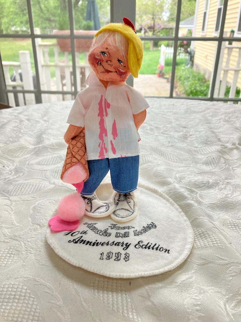May include: A vintage doll with a yellow hat and a white shirt stained with pink. The doll is wearing blue jeans and white sneakers. It is holding a brown ice cream cone and standing on a white base with text that reads "Timothy Annalee Doll Society 10th Anniversary Edition 1993."