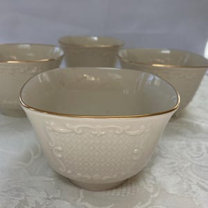 May include: Four off-white ceramic bowls with a square shape and gold trim. The bowls have a decorative embossed pattern. The bowls are arranged on a white tablecloth. The bowls are suitable for serving soup, cereal, or other dishes.