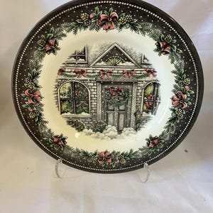 May include: A round ceramic plate featuring a central illustration of a brick house decorated for the holidays with snow, wreaths, and red bows. The border is dark gray with a repeating pattern of green pine branches, red bows, and small pinecones.