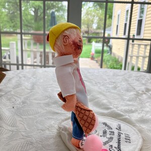 May include: A handmade doll with a yellow cap, white shirt, and blue jeans. The doll is holding a brown object with a pink pom-pom and a pink heart. The doll is standing on a white oval base with text that reads "10th Anniversary Edition 1993".