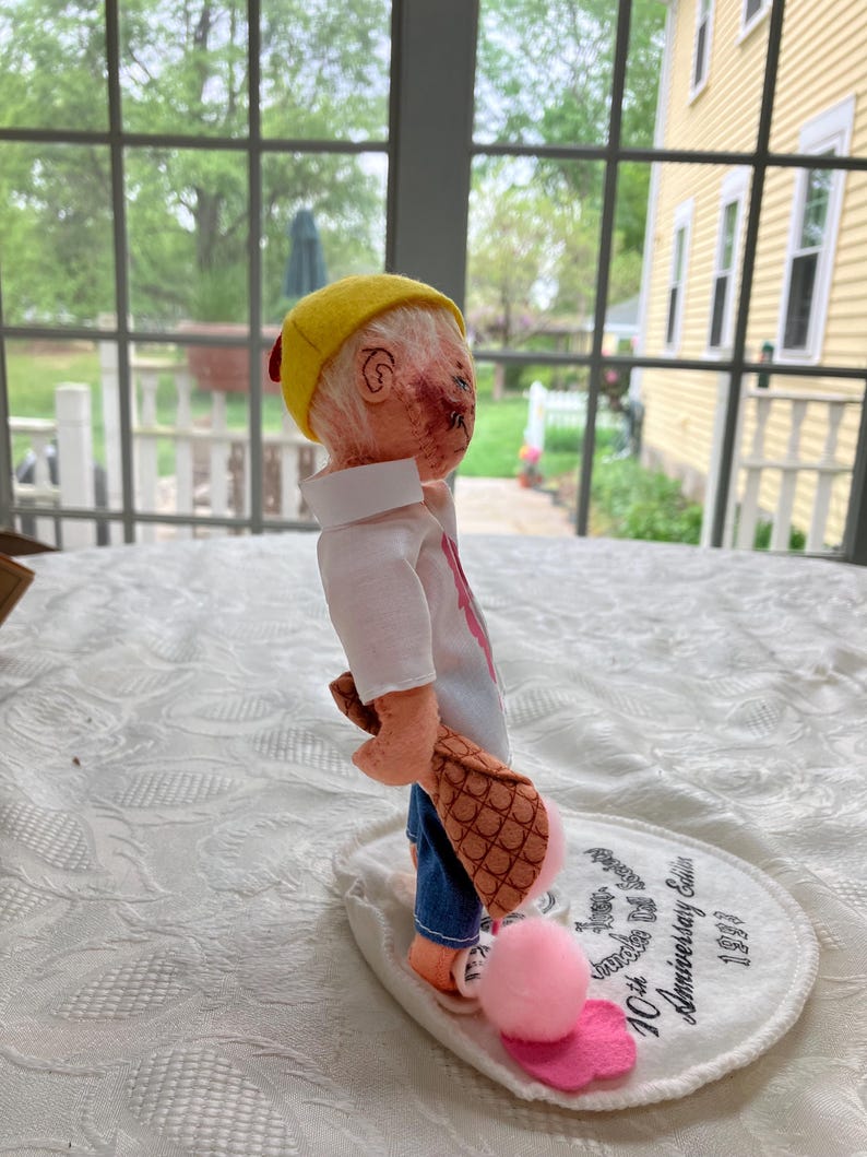 May include: A handmade doll with a yellow cap, white shirt, and blue jeans. The doll is holding a brown object with a pink pom-pom and a pink heart. The doll is standing on a white oval base with text that reads "10th Anniversary Edition 1993".