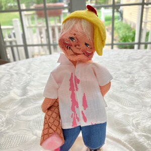 May include: A handmade doll with a whimsical design. The doll wears a yellow hat, a white shirt with pink drips, and blue denim shorts. It holds a brown cone with a pink ice cream scoop and has white shoes with black laces.