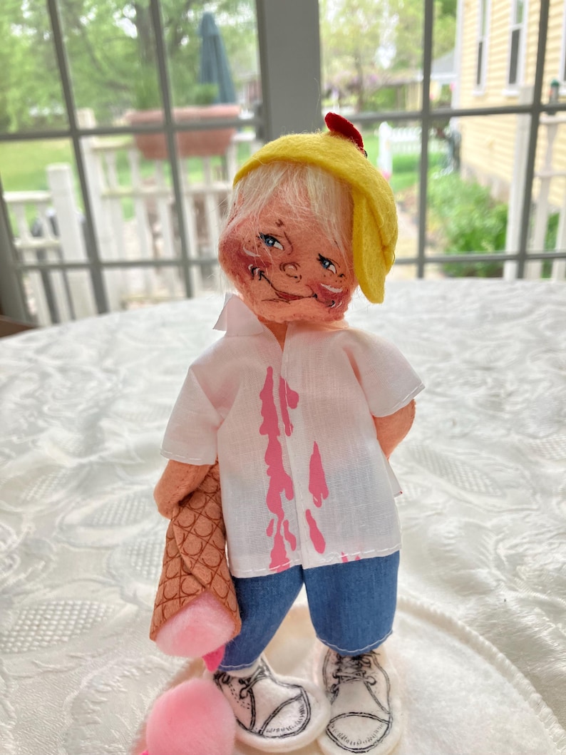 May include: A handmade doll with a whimsical design. The doll wears a yellow hat, a white shirt with pink drips, and blue denim shorts. It holds a brown cone with a pink ice cream scoop and has white shoes with black laces.