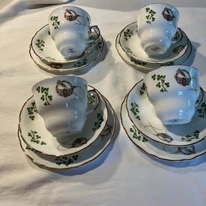 May include: Four white teacups and saucers with gold trim. Each cup and saucer features a green shamrock design and a decorative emblem. The teacups are arranged on a white surface.