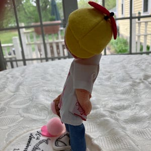 May include: A doll wearing a yellow baseball cap with a red brim, a white shirt, blue jeans, and white sneakers. The doll is holding a pink ice cream cone and a pink ball. The doll is standing on a white circular base with text.
