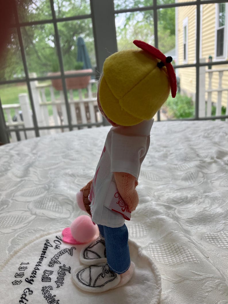 May include: A doll wearing a yellow baseball cap with a red brim, a white shirt, blue jeans, and white sneakers. The doll is holding a pink ice cream cone and a pink ball. The doll is standing on a white circular base with text.