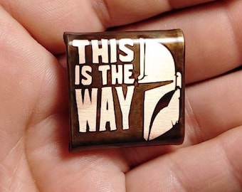 May include: A brown metal pin with the text "This is the Way" and a white silhouette of a Mandalorian helmet.
