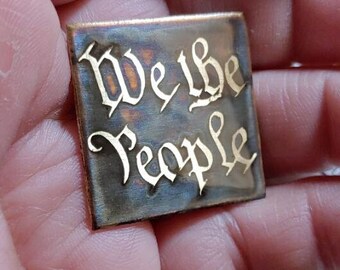 May include: A small, rectangular metal plaque with the words "We the People" engraved in gold lettering. The plaque has a worn, aged look, with a dark blue patina.