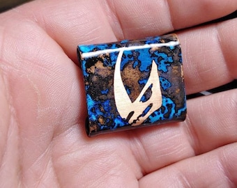 May include: A small, rectangular, copper pendant with a blue and brown marbled finish. The pendant features a white, stylized symbol of a crescent moon.