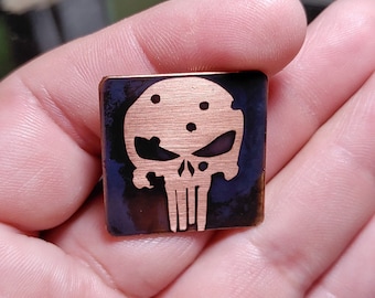 May include: A small, square, copper-colored metal pin with a black background. The pin features a stylized skull with a white outline and a black background. The skull has a small hole in the center of the forehead and two holes on either side of the skull.