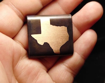 May include: A small, square, metal keychain with a gold outline of the state of Texas. The keychain is dark brown with a brushed metal finish.