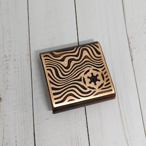 May include: A square wooden coaster with a black and brown wavy pattern and a black Imperial symbol in the centre.