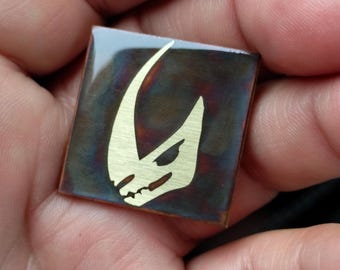 May include: A square-shaped, dark brown, metal pin with a gold-colored Mandalorian symbol on it.