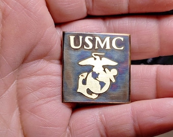 May include: A gold-colored metal pin with the words "USMC" and the Marine Corps emblem.