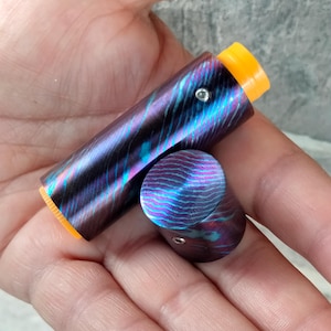 Puede incluir: Un pequeño contenedor cilíndrico de metal morado y azul con una parte superior e inferior de color naranja brillante. El contenedor tiene un patrón de remolino y un pequeño punto plateado en el lateral.