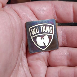 Puede incluir: Un pin de metal cuadrado con acabado negro y dorado. El pin presenta el logotipo de Wu-Tang Clan y el texto "Wu Tang" en letras doradas.
