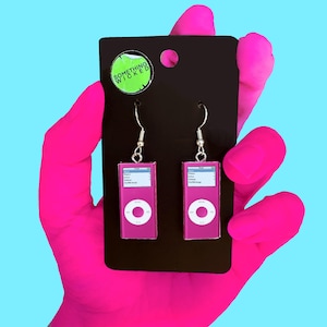 May include: Pink iPod-shaped earrings with white accents and silver hooks. The earrings are displayed on a black card with a green sticker that reads "SOMETHING WICKED". The background is a bright blue.