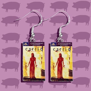May include: A pair of silver earrings with book covers as charms. The book covers are for the novel "Carrie" by Stephen King. The book covers are yellow with a red and white image of a woman in a red dress.