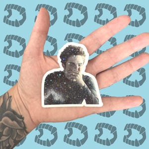 May include: A sticker featuring a portrait of a person with a hand covering their mouth, with a starry design on their shirt. The sticker is held in a hand against a light blue background with repeating vampire teeth.