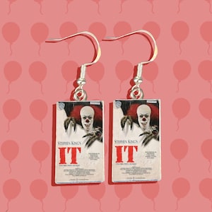 May include: A pair of silver earrings featuring a miniature replica of the Stephen King's IT book cover. The cover features a menacing clown with a red and white costume and a menacing expression. The text on the cover reads "Stephen King's IT".