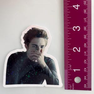 May include: A sticker featuring a portrait of a person with dark hair, covering their mouth with their hand. The sticker is approximately 3 inches tall. The person is wearing a dark long-sleeved shirt with a star pattern.