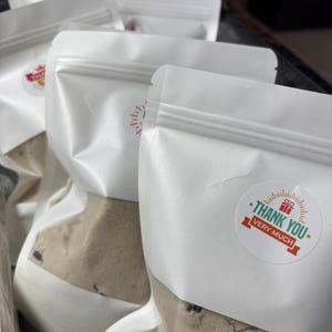 May include: Several white, resealable pouches filled with a light brown mixture, possibly a baking mix. Each pouch has a sticker that says "THANK YOU VERY MUCH" in green and red. The pouches are arranged in a tray.