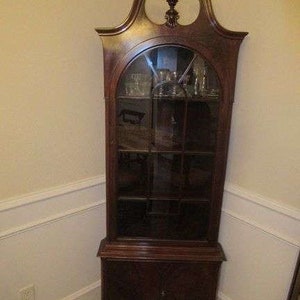 Puede incluir: Un armario de esquina de madera oscura con puertas de vidrio y estantes. El armario tiene una parte superior decorativa con un remate y un armario inferior con una sola puerta.