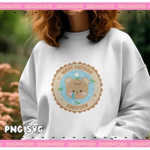 May include: A white sweatshirt with a brown bear illustration wearing a flower crown. The bear is surrounded by a blue circle with the text "Went Outside Survivor" in brown lettering.