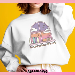 May include: White sweatshirt with a colourful illustration of a window scene. The design includes books, a pumpkin, and fairy lights. The window shows a sunset sky with stars and clouds. The text "ABCOMICSVG" is at the bottom.