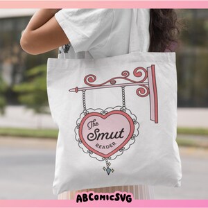 May include: White tote bag with a pink heart-shaped sign that says "The Smut Reader".