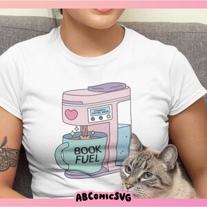 May include: White t-shirt featuring a pink coffee maker with a heart and the text "STRONK ENTIRE BOOK". A blue mug with "BOOK FUEL" is under the coffee maker. A cat with blue eyes is in the image.