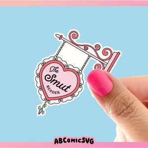 May include: A pink heart-shaped sign with the text "The Smut Reader" hanging from a decorative metal frame. The sign is on a light blue background.