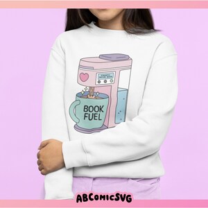 May include: White sweatshirt featuring a pastel pink coffee maker graphic with a heart and the text "BOOK FUEL" on a teal mug. The coffee maker is pouring coffee into the mug. The sweatshirt is a comfortable, casual style.