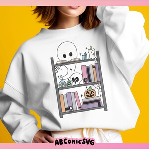 May include: A white sweatshirt featuring a graphic of a ghost reading a book on a bookshelf with Halloween decorations. The decorations include a pumpkin, a skull, a candle, and potion bottles.