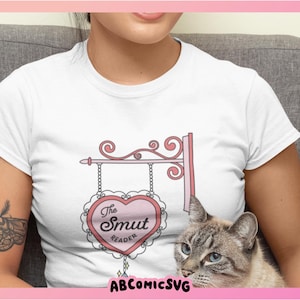 May include: White t-shirt with a pink heart-shaped sign that says "The Smut Reader" on it. The sign is hanging from a decorative metal frame.