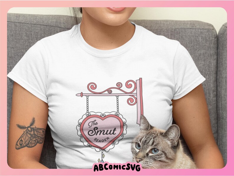 May include: White t-shirt with a pink heart-shaped sign that says "The Smut Reader" on it. The sign is hanging from a decorative metal frame.