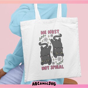 May include: White tote bag featuring a cartoon design of two gray cats, flowers, and the text "WE MUST NOT SPIRAL" in pink. The bag has white handles and is held by a person wearing a light blue sweater.