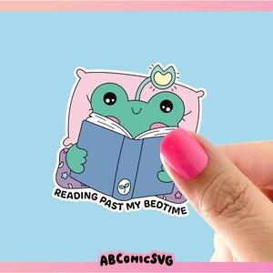 Op de afbeelding: Een groene kikker die een roze kussen en een lichtblauw boek met een groen blaadjesontwerp draagt. De kikker leest het boek en de tekst zegt "Reading past my bedtime".