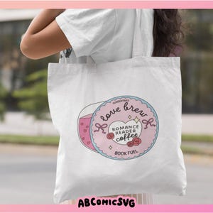 Puede incluir: Bolso de mano blanco con un gr&aacute;fico circular con las palabras "Love Brew" y "Romance Reader Coffee". El dise&ntilde;o incluye una taza de caf&eacute; rosa, corazones y rosas. Tambi&eacute;n est&aacute; presente el texto "Book Fuel". La bolsa tiene el texto "ABCOMICSVG" en la parte inferior.